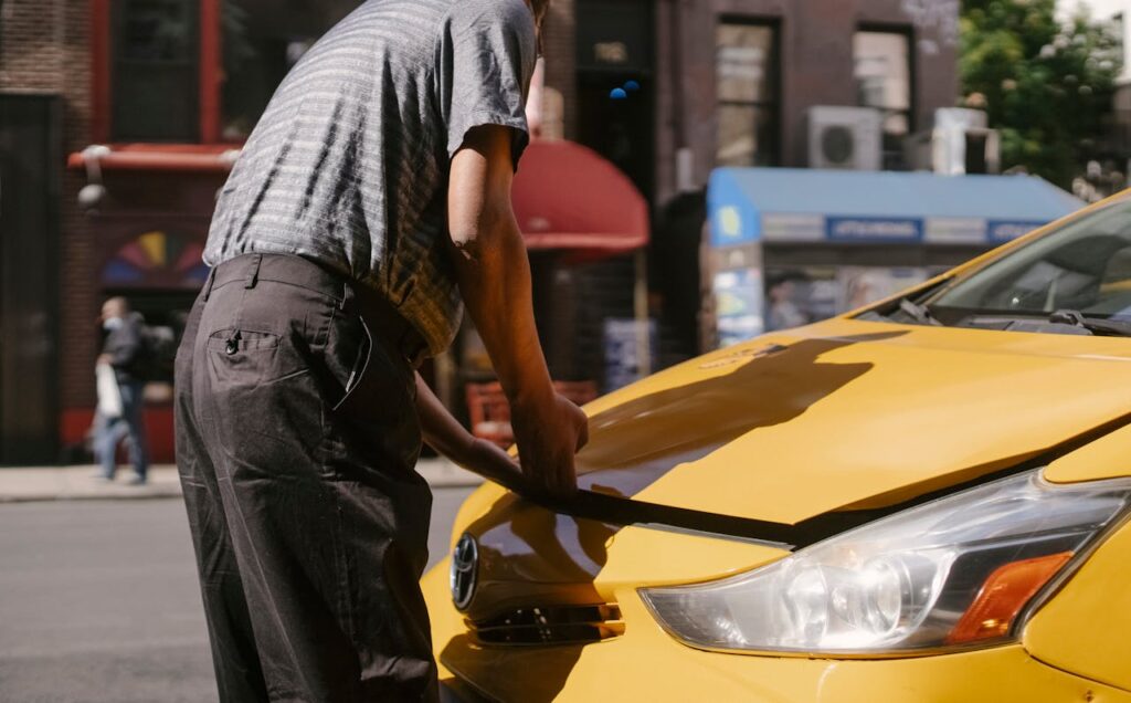 pexels-photo-5835351 Man inspecting open hood of a yellow taxi on city street.